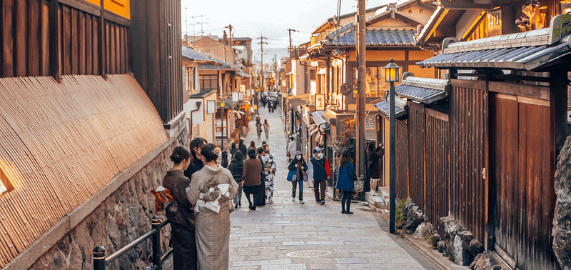 Personas caminando por una calle de Kioto bordeada de edificios antiguos al atardecer.