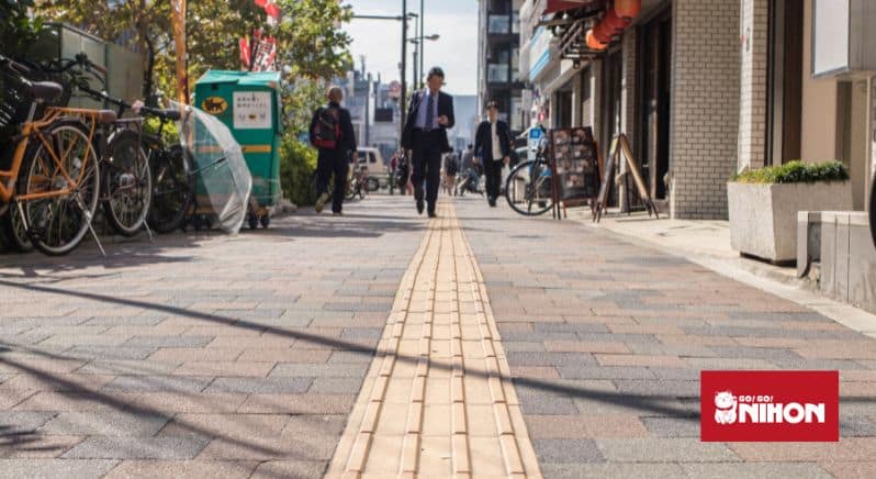 Imagen a pie de calle que muestra un pavimento táctil amarillo con gente caminando al fondo