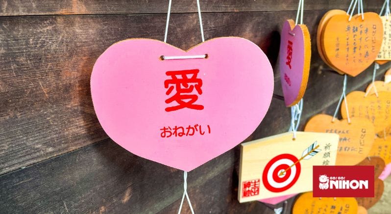 Bild einer rosafarbenen Ema-Tafel, die in einem Tempel in Japan hängt, mit dem Zeichen für Liebe (愛) und おねがい (bitte)