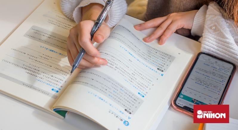 Jemand hält einen Stift auf einem JLPT N4 Lernbuch neben einem Telefon. 