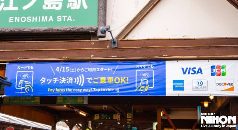 En banner hänger över ingången till Enoshima station med information kring kreditkortbetalning för tågresor. 