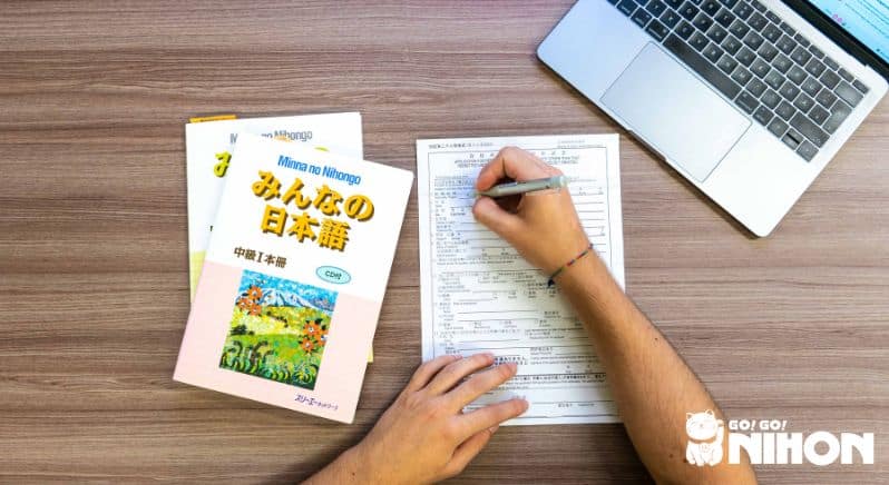 bild ovanfrån på två händer som fyller i ett formulär med japanska läroböcker till vänster om sig och en bärbar dator till höger.
